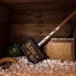 A wooden crate interior, showcasing Gallagher&rsquo;s iconic Sledge-O-Matic hammer resting upright among scattered packing peanuts and slightly overripe bananas.