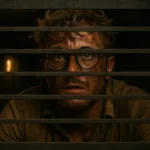 A disheveled man with messy blonde hair, sweat on his brow, and dirt-smudged clothes peering through a metal vent grate from inside a dusty air duct.