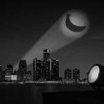 A dramatic nighttime cityscape of Detroit seen from a suburban rooftop, where a spotlight projects a glowing banana-shaped signal into the cloudy sky, mimicking the iconic Bat-Signal.