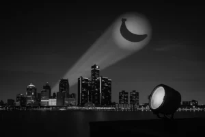 A dramatic nighttime cityscape of Detroit seen from a suburban rooftop, where a spotlight projects a glowing banana-shaped signal into the cloudy sky, mimicking the iconic Bat-Signal.